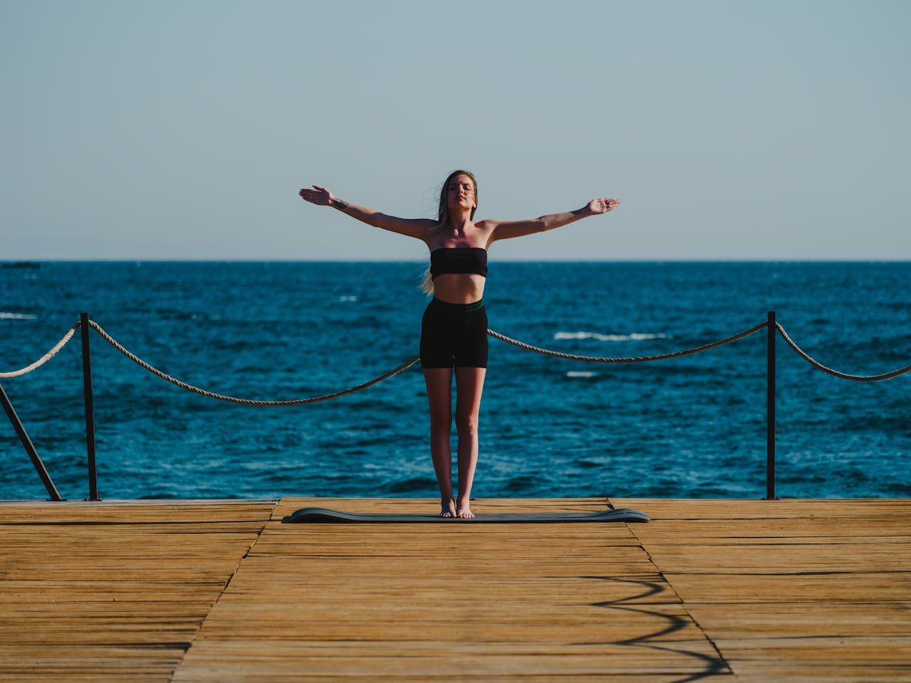 A woman in activewear stands in a yoga pose on a wooden dock near the sea, embracing a tranquil moment.